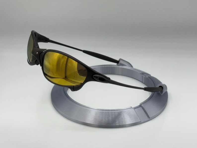 May include: Black sunglasses with gold-tinted lenses rest on a gray circular display stand. The sunglasses have a sleek, modern design with a black frame and arms. The lenses reflect a golden hue.