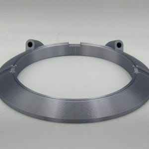 May include: A gray, circular object with a flat, beveled edge and two raised sections with holes. The object has a metallic appearance and is set against a white background. The design is modern and minimalist.