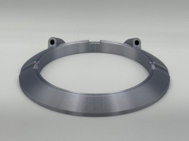 May include: A gray, circular object with a flat, beveled edge and two raised sections with holes. The object has a metallic appearance and is set against a white background. The design is modern and minimalist.