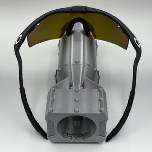 May include: Black sunglasses with yellow-tinted lenses rest on top of a gray, 3D-printed bomb-shaped object. The sunglasses have a curved design with black frames and arms. The bomb has a cylindrical body and a detailed, textured surface.