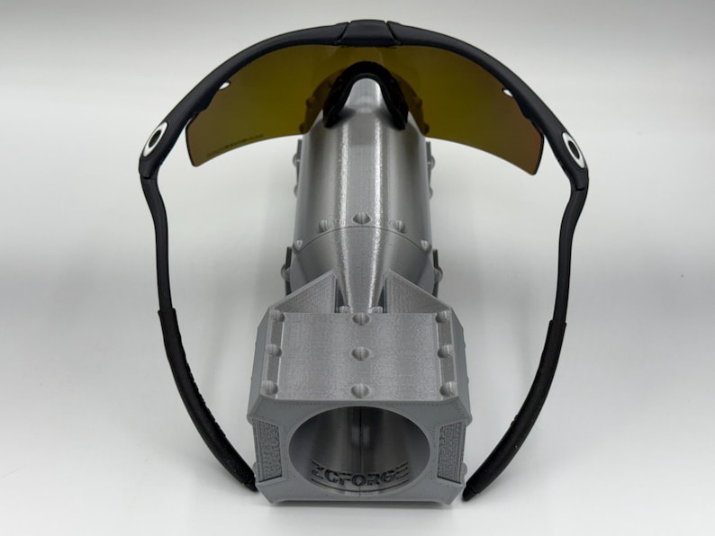 May include: Black sunglasses with yellow-tinted lenses rest on top of a gray, 3D-printed bomb-shaped object. The sunglasses have a curved design with black frames and arms. The bomb has a cylindrical body and a detailed, textured surface.
