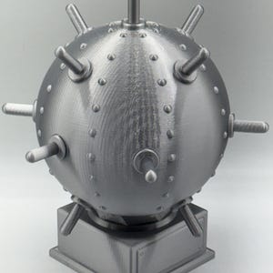 May include: A grey, spherical object featuring protruding cylindrical elements and small, rounded studs. It is mounted on a square base with a rectangular plaque. The design suggests a mechanical or industrial theme.