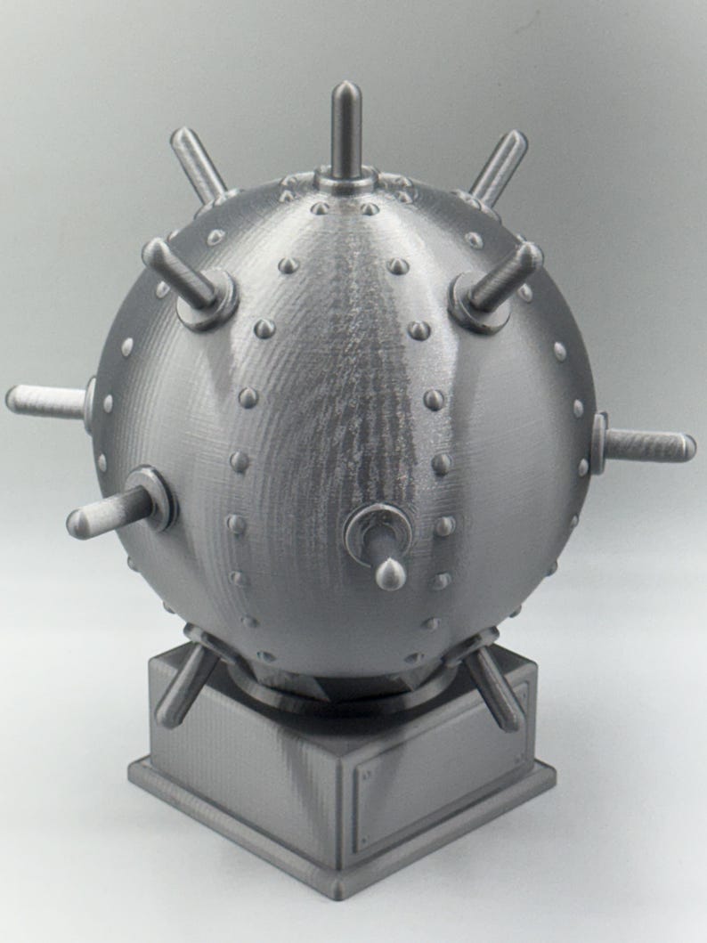 May include: A grey, spherical object featuring protruding cylindrical elements and small, rounded studs. It is mounted on a square base with a rectangular plaque. The design suggests a mechanical or industrial theme.