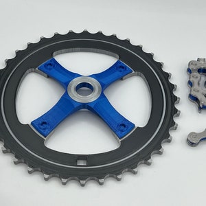 Pu&ograve; includere: Un pignone per bicicletta nero e argento con un design a forma di croce blu al centro. Sono mostrati anche due anelli di catena separati.