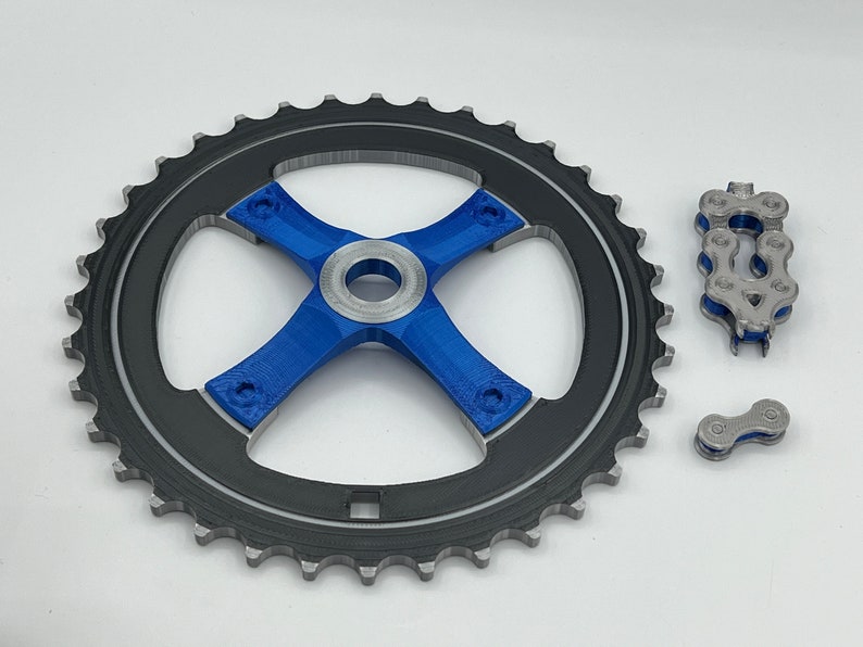 Pu&ograve; includere: Un pignone per bicicletta nero e argento con un design a forma di croce blu al centro. Sono mostrati anche due anelli di catena separati.