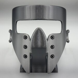 May include: A gray, 3D-printed mask with a distinctive design. The mask features a prominent, pointed central piece and is secured with visible screws. It has a wide opening for the eyes and is designed to fit the face.