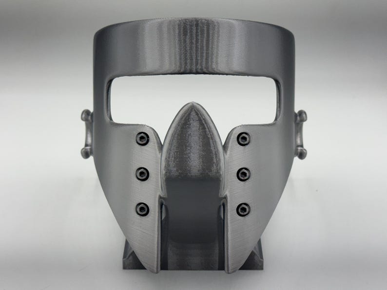 May include: A gray, 3D-printed mask with a distinctive design. The mask features a prominent, pointed central piece and is secured with visible screws. It has a wide opening for the eyes and is designed to fit the face.