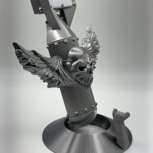 May include: A gray, 3D-printed sculpture of a bomb with a skull and wings emblem. The bomb has a cylindrical body, a square top, and is mounted on a circular base. The sculpture is detailed with rivets and a support piece.