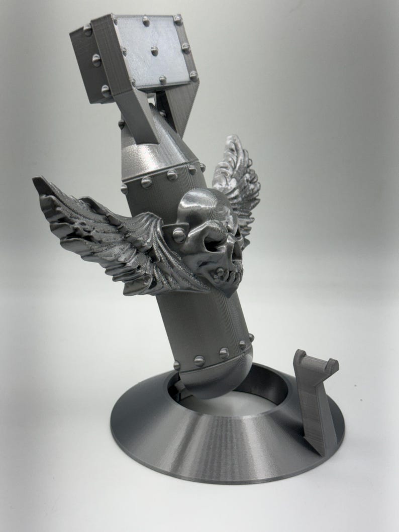 May include: A gray, 3D-printed sculpture of a bomb with a skull and wings emblem. The bomb has a cylindrical body, a square top, and is mounted on a circular base. The sculpture is detailed with rivets and a support piece.