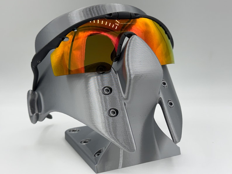 May include: Black sports sunglasses with orange-yellow lenses rest on a gray display stand. The stand has a unique, angular design with multiple screw details. The lenses reflect a warm, vibrant glow.