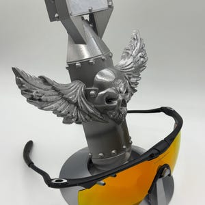 May include: A silver-colored bomb-shaped sunglasses holder with a skull and wing design. The holder supports a pair of black-framed sunglasses with orange lenses. The sunglasses are displayed on a round base.