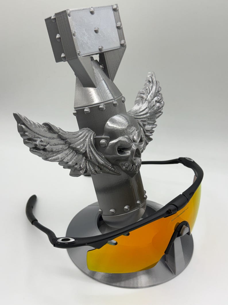 May include: A silver-colored bomb-shaped sunglasses holder with a skull and wing design. The holder supports a pair of black-framed sunglasses with orange lenses. The sunglasses are displayed on a round base.