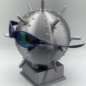 May include: A silver spherical sculpture with spikes, similar to a mine, displays blue-framed sunglasses with iridescent lenses. The sculpture rests on a square base, creating a unique display. The sculpture is approximately 15 cm in diameter.