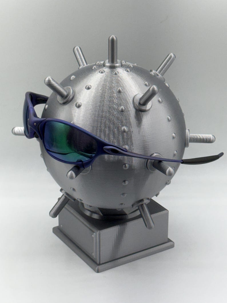 May include: A silver spherical sculpture with spikes, similar to a mine, displays blue-framed sunglasses with iridescent lenses. The sculpture rests on a square base, creating a unique display. The sculpture is approximately 15 cm in diameter.