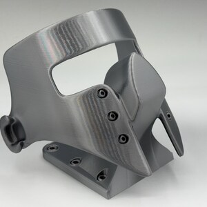 May include: A gray, 3D-printed mask with a curved design. The mask features a prominent nose piece and is secured with small screws. It has a base with multiple holes, suggesting it may be mounted or attached to something.