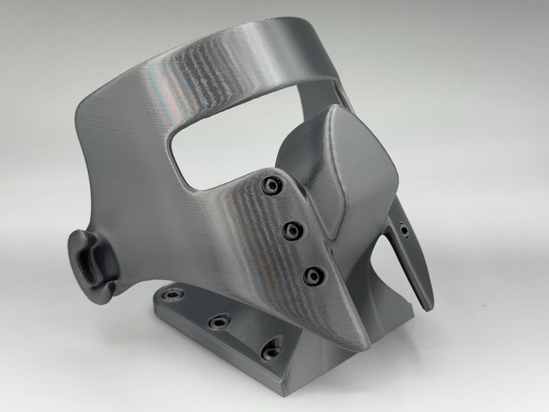 May include: A gray, 3D-printed mask with a curved design. The mask features a prominent nose piece and is secured with small screws. It has a base with multiple holes, suggesting it may be mounted or attached to something.