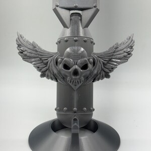 May include: A gray, 3D-printed decorative bomb with a skull and wings design. The bomb features a cylindrical body, a rectangular top, and a circular base. The skull is centered on the body with detailed wings extending outward.
