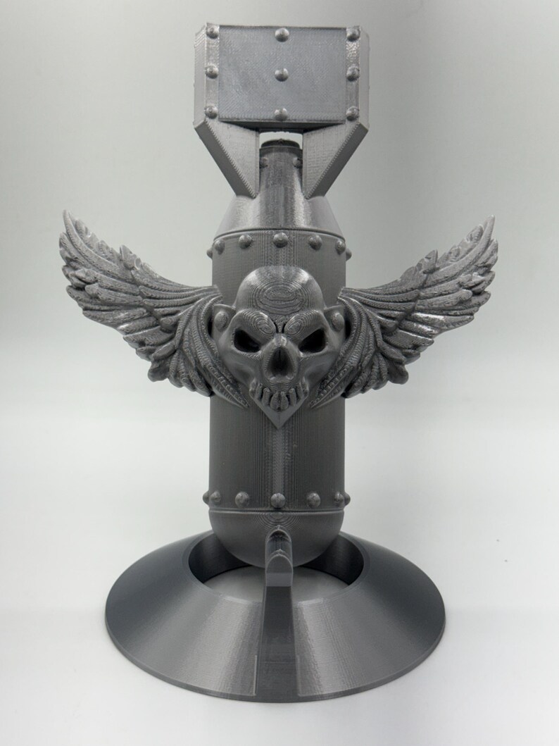 May include: A gray, 3D-printed decorative bomb with a skull and wings design. The bomb features a cylindrical body, a rectangular top, and a circular base. The skull is centered on the body with detailed wings extending outward.