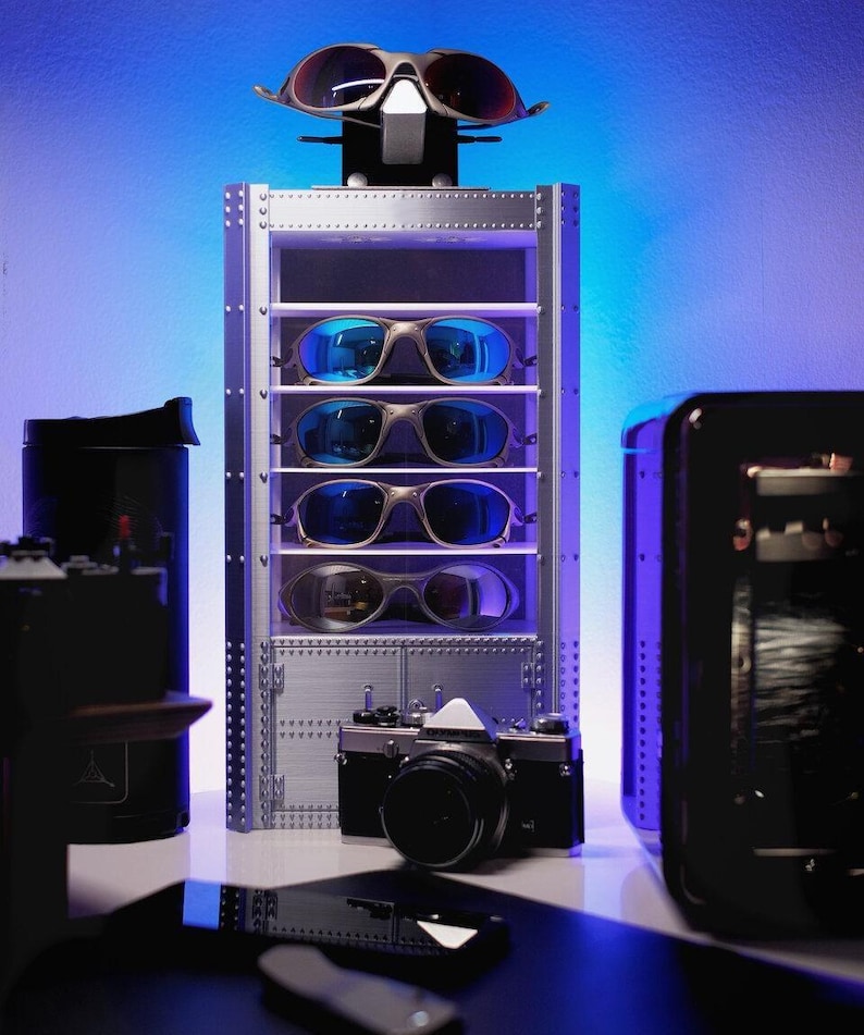K&ouml;nnte beinhalten: Eine silberne Vitrine mit mehreren Sonnenbrillen mit blauen und verspiegelten Gl&auml;sern. Vorne steht eine Vintage-Kamera. Der Hintergrund hat einen blauen und violetten Farbverlauf.