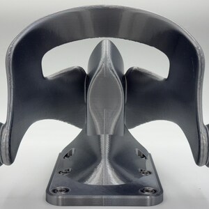 May include: A gray, 3D-printed bracket with a curved top and a base with screw holes. The bracket has a symmetrical design with rounded edges and a textured surface. The overall shape resembles a stylized mask or face.