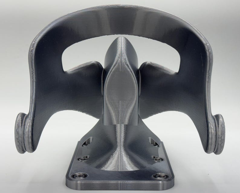 May include: A gray, 3D-printed bracket with a curved top and a base with screw holes. The bracket has a symmetrical design with rounded edges and a textured surface. The overall shape resembles a stylized mask or face.