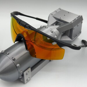 May include: Black-framed sunglasses with orange-tinted lenses rest on a gray, metallic, industrial-style sculpture. The sculpture has a cylindrical base and a boxy top, with visible rivets and a matte finish.