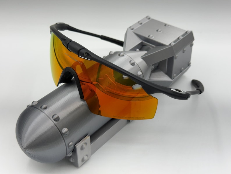 May include: Black-framed sunglasses with orange-tinted lenses rest on a gray, metallic, industrial-style sculpture. The sculpture has a cylindrical base and a boxy top, with visible rivets and a matte finish.