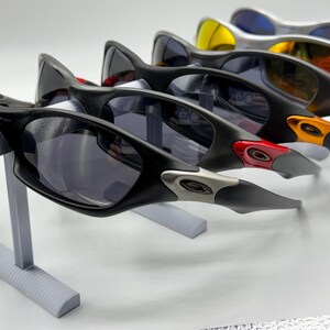 May include: A display of several pairs of sunglasses with black frames and tinted lenses. The sunglasses are arranged on a gray stand, showcasing their design and color variations, including red, orange, and yellow accents.