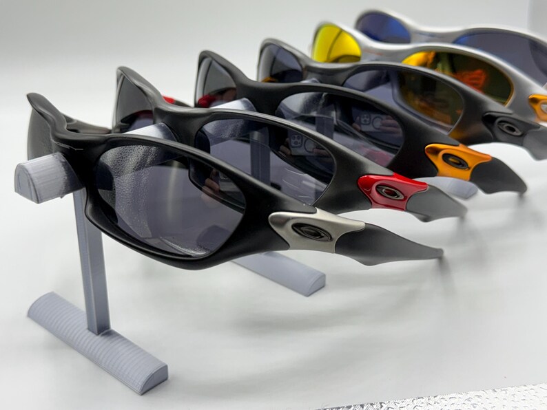 May include: A display of several pairs of sunglasses with black frames and tinted lenses. The sunglasses are arranged on a gray stand, showcasing their design and color variations, including red, orange, and yellow accents.