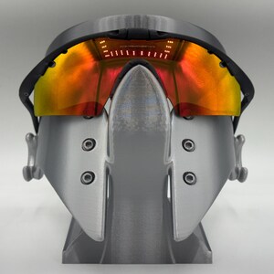 May include: A pair of futuristic-looking sunglasses with a black frame and orange-yellow lenses. The glasses are mounted on a gray stand. The lenses have a gradient effect, transitioning from orange to yellow. The stand is also gray.