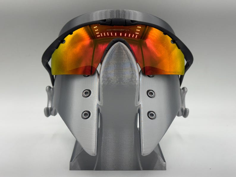 May include: A pair of futuristic-looking sunglasses with a black frame and orange-yellow lenses. The glasses are mounted on a gray stand. The lenses have a gradient effect, transitioning from orange to yellow. The stand is also gray.