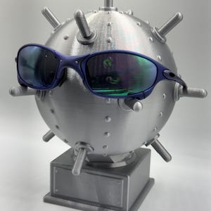 May include: Blue-framed sunglasses with reflective lenses are positioned on a metallic, spherical sculpture, similar to a mine. The sculpture features protruding cylindrical elements and is mounted on a rectangular base.