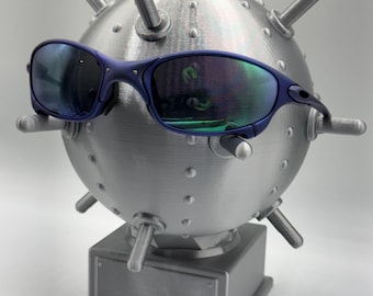 May include: A pair of blue-framed sunglasses with reflective lenses rests on a metallic, spherical sculpture resembling a mine. The sculpture has protruding cylindrical elements and sits on a rectangular base.