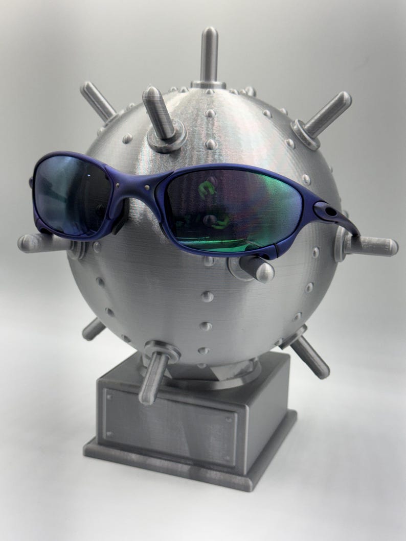 May include: Blue-framed sunglasses with reflective lenses are positioned on a metallic, spherical sculpture, similar to a mine. The sculpture features protruding cylindrical elements and is mounted on a rectangular base.