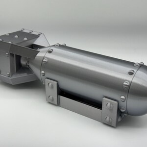 May include: A gray, 3D-printed model of a bomb. The model has a cylindrical body with a rounded end and a rectangular tail section. The surface is detailed with rivets and mounting brackets.