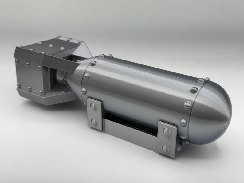 May include: A gray, 3D-printed model of a bomb. The model has a cylindrical body with a rounded end and a rectangular tail section. The surface is detailed with rivets and mounting brackets.
