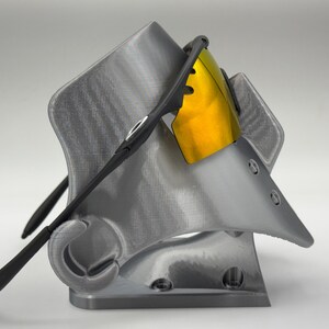 May include: A gray 3D-printed sunglasses stand with a pair of black sunglasses with yellow lenses. The stand has a unique, angular design with a curved hook for the glasses' arms.