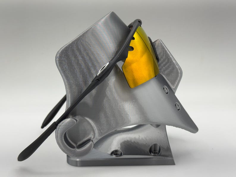 May include: A gray 3D-printed sunglasses stand with a pair of black sunglasses with yellow lenses. The stand has a unique, angular design with a curved hook for the glasses' arms.