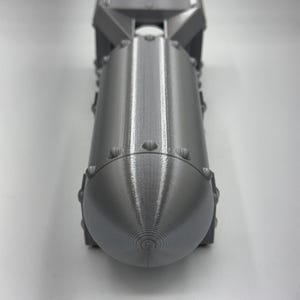 May include: A gray, 3D-printed model of a bomb. The model has a cylindrical body with a rounded nose and a fin assembly at the top. The surface has a textured appearance with visible layer lines from the printing process.