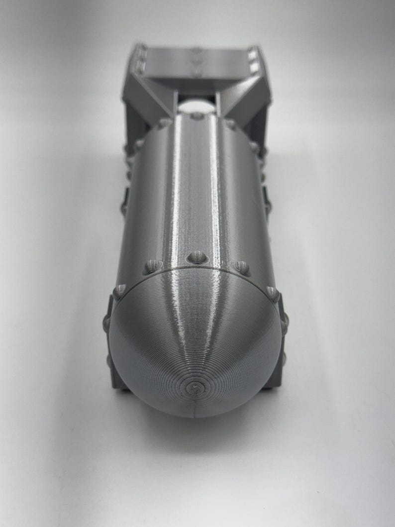 May include: A gray, 3D-printed model of a bomb. The model has a cylindrical body with a rounded nose and a fin assembly at the top. The surface has a textured appearance with visible layer lines from the printing process.