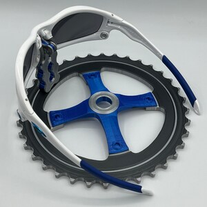 Pu&ograve; includere: Occhiali da sole bianchi e blu con lente grigio scuro, piegati e appoggiati su un pignone di bicicletta nero e argento.