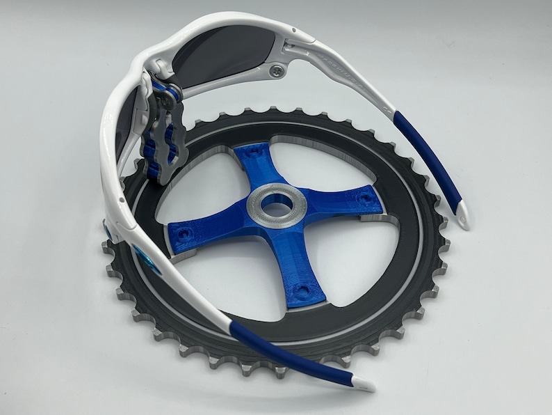 Pu&ograve; includere: Occhiali da sole bianchi e blu con lente grigio scuro, piegati e appoggiati su un pignone di bicicletta nero e argento.
