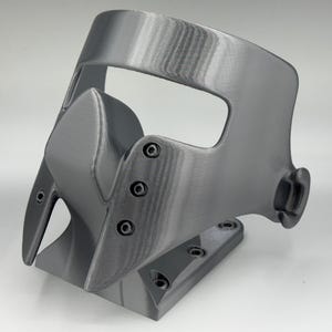 May include: A gray, 3D-printed mask with a curved design. The mask features a nose piece, eye openings, and a base with screw holes. The surface has visible layer lines from the printing process.
