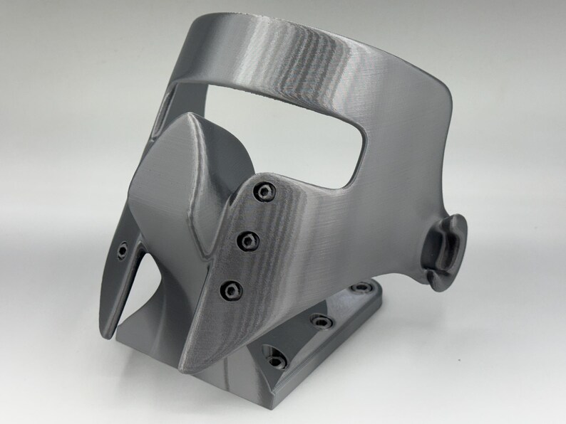 May include: A gray, 3D-printed mask with a curved design. The mask features a nose piece, eye openings, and a base with screw holes. The surface has visible layer lines from the printing process.