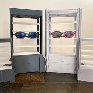 K&ouml;nnte beinhalten: Ausstellungsregale in verschiedenen Gr&ouml;&szlig;en, grau und wei&szlig; lackiert, mit mehreren Regalb&ouml;den und Schrankt&uuml;ren. Zwei Sonnenbrillen mit blauen und roten Gl&auml;sern werden pr&auml;sentiert. Die Regale sind f&uuml;r die Einzelhandelspr&auml;sentation konzipiert.