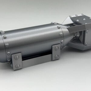 May include: A gray, metallic model of a bomb. The cylindrical body has a rounded nose and is secured with rivets. The tail section is a rectangular shape with a fin. A handle is attached to the bottom.