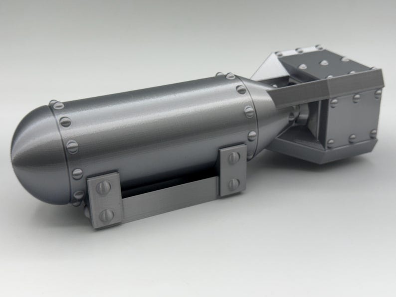 May include: A gray, metallic model of a bomb. The cylindrical body has a rounded nose and is secured with rivets. The tail section is a rectangular shape with a fin. A handle is attached to the bottom.