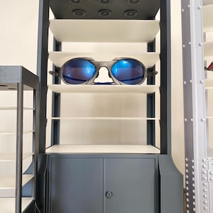K&ouml;nnte beinhalten: Ein grauer Display-Schrank mit mehreren wei&szlig;en Regalen und einem Unterschrank. Eine Sonnenbrille mit blauen Gl&auml;sern liegt auf einem der Regale. Der Schrank hat einen dunkelgrauen Rahmen und kreisf&ouml;rmige Akzente auf der Oberseite.