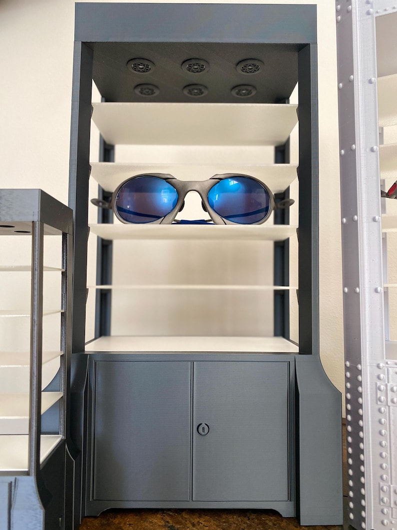 K&ouml;nnte beinhalten: Ein grauer Display-Schrank mit mehreren wei&szlig;en Regalen und einem Unterschrank. Eine Sonnenbrille mit blauen Gl&auml;sern liegt auf einem der Regale. Der Schrank hat einen dunkelgrauen Rahmen und kreisf&ouml;rmige Akzente auf der Oberseite.