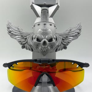 May include: Black-framed sunglasses with orange-red lenses rest on a gray, bomb-shaped display stand. The stand features a skull with wings. The sunglasses are the focal point, showcasing their vibrant lenses and sleek design.